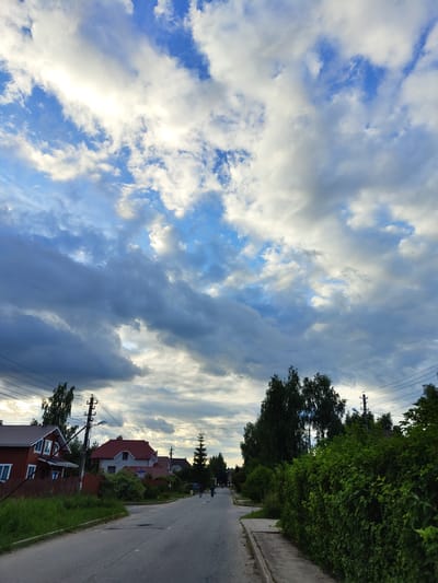 The image depicts a street scene in a residential area, likely in Цибино, Russia. The perspective is from a low angle looking upwards and across the street. Dominating the left side of the frame is a dramatic sky filled with a mix of bright white and dark gray clouds against a blue sky. The clouds are textured and appear to be moving, suggesting a dynamic weather pattern, possibly with sunshine breaking through.
On the right side of the image, a paved road runs into the distance, lined with a sidewalk and lush green trees and hedges. Several houses with red roofs are visible along the street, interspersed with trees and utility poles. The architecture suggests a suburban or semi-rural setting. Two figures, possibly pedestrians, are visible walking on the road in the distance.
The time of day appears to be late afternoon or early evening, given the quality of the light and the dramatic sky. The presence of utility poles and overhead wires indicates that electricity is supplied to the area. The overall atmosphere is one of quiet everyday life in a village or small town. There is no visible text in the image.