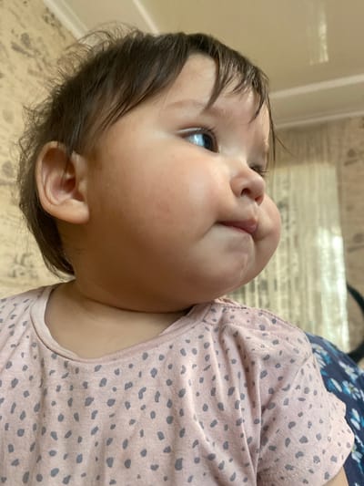 The image shows a close-up, sideways view of a baby's face and upper body. The baby has dark hair, blue eyes, and chubby cheeks. They are wearing a light pink shirt with a pattern of greyish-blue polka dots. The background is blurred but appears to be an indoor setting with a light-colored wall and floor. The baby's head is tilted upwards and to the side, and they seem to be looking at something or someone off-camera. The lighting suggests it is daytime. The overall mood is calm and intimate, capturing a candid moment with the baby. There is no visible text in the image.