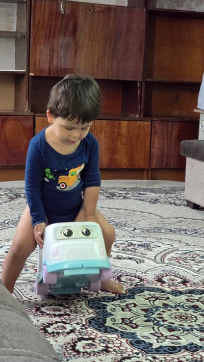 A young child, appearing to be between two and three years old, is seated on a toy vehicle indoors. The child is wearing a dark blue long-sleeved bodysuit with a graphic of a yellow truck with green accents. The child's legs are visible, extending downwards from the toy. The toy vehicle is white and light blue with large, cartoonish eyes and pink wheels. The child is looking down at the toy, and their hands are positioned on either side of it. The setting is an indoor environment with a patterned carpet covering the floor. In the background, there is a large, dark wooden cabinet with multiple shelves. This scene is located in the city of Yakkabog, Uzbekistan.