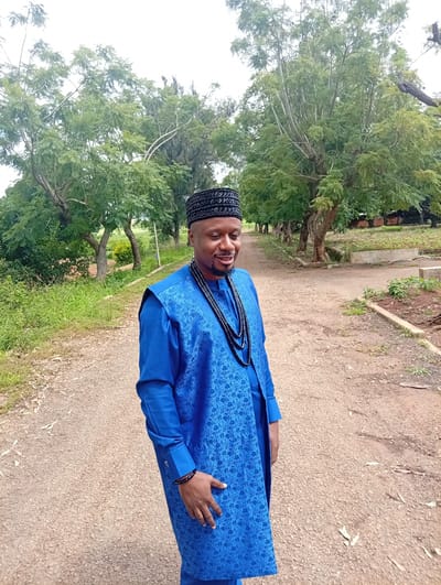 A man wearing a blue traditional outfit and a black patterned cap stands on a dirt path in a grassy area with trees. He is smiling and looking down to his right. The outfit consists of a long-sleeved blue shirt and a blue vest with a patterned design. He is also wearing a black beaded necklace and a black beaded bracelet on his left wrist. The path appears to be in a rural or suburban setting, possibly a park or a residential area. The background shows lush green trees and vegetation. The location is specified as Tambes Mile Four, Nigeria.