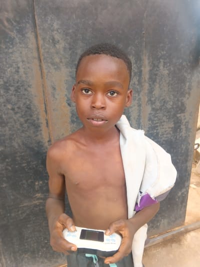 A young male, approximately 8-12 years old, with dark skin and short dark hair, stands shirtless, facing the viewer with a neutral expression. He wears dark-colored shorts with a light accent stripe. A light-colored garment, possibly a shirt, is draped over his left shoulder. Both hands hold a white handheld electronic gaming device with a central screen and light blue buttons, labeled "GAME BOX." The background consists of a dark, weathered, possibly metallic panel or gate, showing signs of rust and discoloration. A light brown ground surface is visible at the lower right. The scene is located in Itu, Nigeria.