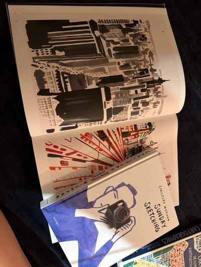 The image shows an open book with ink sketches on both pages. The left page features a black and gray abstract ink drawing of what appears to be a cityscape or architectural elements. The right page contains a vibrant ink sketch with red and green lines radiating from a central point, possibly representing a stylized explosion or burst. Resting on the open book is a hardcover book titled "SUNDAY SKETCHING" by CHRISTOPH NIEMANN, which has a cover illustration of a person holding a camera. Another book titled "CARTOGRAPHICS" with the subtitle "DIE KUNST DER KARTENGESTALTUNG" is visible to the right, displaying a colorful map-like illustration. The books and the open sketchbook are placed on a dark, soft surface, likely fabric, casting shadows. There is no indication of the city of Mühlbach - Rio di Pusteria, Italy within the image.