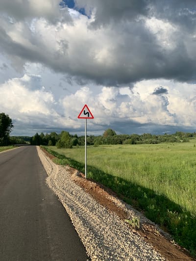 This is a photograph of a rural road in the city of Svyatitsa, Russia. The road is paved and curves to the left, with a gravel shoulder on the right. A traffic sign warning of sharp curves ahead is visible on the right side of the road. The surrounding landscape is a green field with trees in the distance. The sky is filled with dramatic, dark clouds.