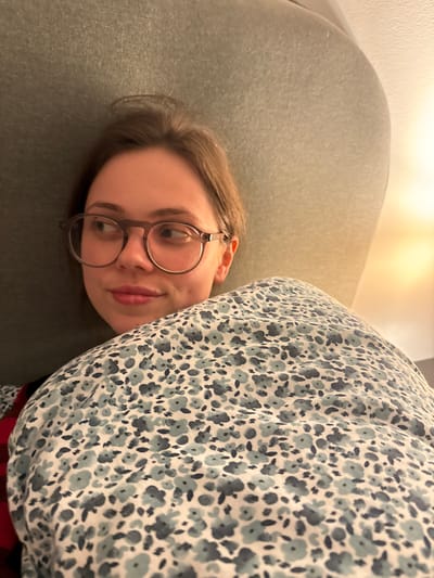 A young woman lies on a gray upholstered surface, likely a couch or bed, partially covered by a light-colored floral patterned blanket. She wears round-framed glasses and gazes towards the camera with a slight smile. The setting appears to be indoors, possibly a home. There are no discernible details connecting the image to Perugia, Italy.