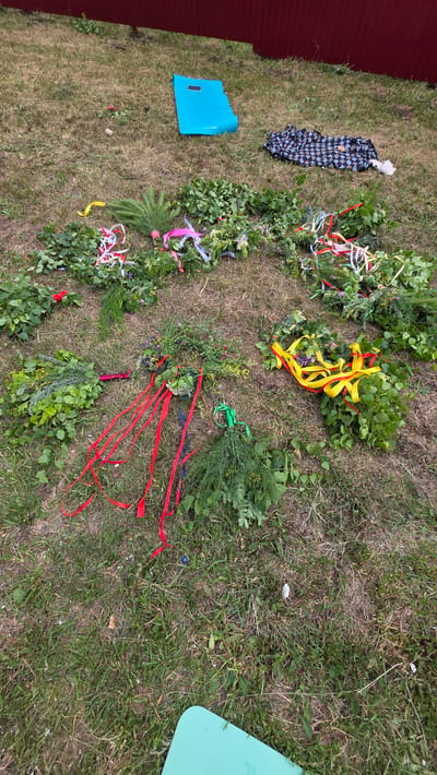 The image depicts an outdoor scene on a grassy area, likely in the city of Kupoyarovo, Russia. The ground is covered in dry grass and scattered debris. In the foreground, several arrangements of greenery and colorful ribbons are laid out on the grass. These arrangements appear to be bouquets or wreaths made of leaves, flowers, and possibly branches. Ribbons of various colors, including red, yellow, white, and green, are tied to the greenery.
In the middle ground, a bright blue mat or yoga mat is placed on the grass. To its right, a folded dark blue and white checkered blanket is visible. Further back, a red wooden fence or wall runs along the top of the image, partially obscured by the overgrowth. The lighting suggests it is daytime, possibly overcast or with soft sunlight. No people are visible in the image.