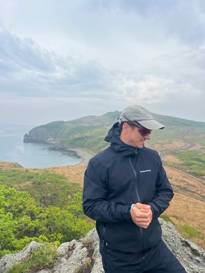 A man in a dark jacket, light-colored cap, and sunglasses is standing on a rocky outcrop. He is looking downwards and slightly to his left. The landscape behind him features a hillside with a mix of green vegetation and brown, grassy areas. In the distance, there is a body of water and a cloudy sky. The location is Gvozdevo, Russia.