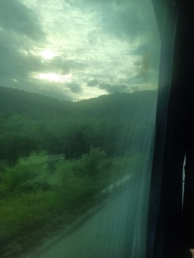 The image is a scenic view from a window, likely from a vehicle, overlooking a lush green landscape of trees and hills under a cloudy sky. The sun is visible through the clouds, casting a soft light. There are power lines stretching across the sky and a sign is affixed to the glass of the window. The overall impression is one of a journey through a natural, perhaps rural, setting. The location in Kamianytsia, Ukraine, suggests a picturesque area with natural beauty.
