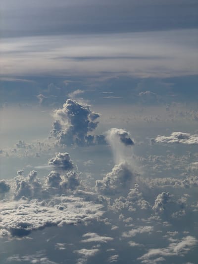 The image shows an aerial view of clouds. The sky is a mixture of blue and gray. Various cloud formations are visible, including cumulus and cirrus clouds. The image does not depict any specific location or city. It does not contain any people, subjects, or objects other than clouds. No actions or interactions are visible. The environment is the atmosphere.