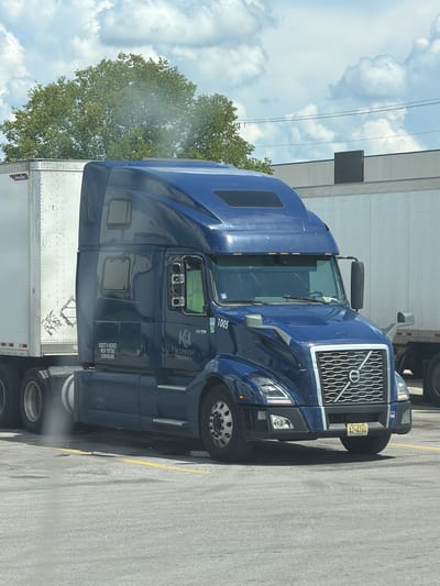 A dark blue Volvo semi-truck, labeled "K&L EXPRESS LOGISTICS" and numbered "1005", is parked in a paved lot in Lebanon, United States of America. The truck has a white trailer attached and is positioned in front of a light-colored building with a dark rectangular panel. The sky is partly cloudy, with green trees visible in the background. A yellow line runs across the pavement in front of the truck. The truck's license plate reads "AZ 4760".