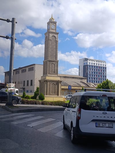 The image shows a street scene in the Bayrampaşa district of Istanbul, Turkey. In the foreground, a white Ford Tourneo van is parked on the right side of the road, with its rear license plate clearly visible: "06 EZT 223". A pedestrian crossing is in the foreground. To the left, a black car is parked. In the middle ground, a tall, ornate clock tower stands prominently, adorned with intricate geometric patterns. Behind the clock tower, a long, low building with a light-colored facade is visible. Further in the background, a modern, multi-story building with a dark facade and red-trimmed windows stands out, identified by a sign as "Ramada Encore". The sky is blue with scattered white clouds. The scene appears to be a typical urban environment on a bright day, with a mix of historical and modern architecture.