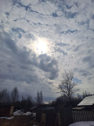The image depicts a rural scene in Avdot’ino, Russia, under a partly cloudy sky. The sun is visible in the upper-left quadrant, partially obscured by clouds, casting a bright glare. The sky features scattered cumulus and stratus clouds, with some patches of visible blue. In the foreground, the ground consists of bare, dark earth interspersed with patches of melting snow. Several rustic structures are visible; to the left, a reddish-brown building with a light-colored door frame, and on the right, a building featuring a corrugated metal roof. A dark wooden fence extends across the midground. Numerous deciduous trees, bare of leaves, are present throughout the scene, with a prominent, larger leafless tree silhouetted against the sky in the right midground. A dense treeline forms the distant background. Three parallel overhead utility lines traverse diagonally from the upper-left to the upper-right of the frame. A utility pole with a crossarm is discernible in the distant center-left. No individuals or animals are visible, and no discernible actions are occurring.
