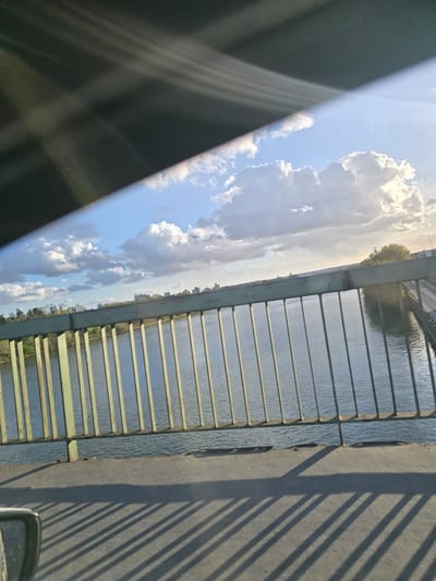 The image depicts a panoramic view from a vehicle positioned on a bridge over a body of water, likely the Seine River, in Argenteuil, France. The upper left quadrant is obscured by a dark, angled structure, consistent with a vehicle's interior roof or sun visor, exhibiting linear light streaks. The sky, visible in the upper and central portions, is bright blue with scattered white cumulus clouds, and direct sunlight creates a prominent lens flare in the upper right.

A green-painted metal bridge railing, consisting of evenly spaced vertical bars, extends horizontally across the mid-ground. Beneath this railing, the river's surface displays subtle ripples, reflecting the sky. The distant left bank features a low horizon line of green trees and a series of multi-story buildings. To the right, an illuminated landmass with dense foliage is visible, along with a receding section of a road or another bridge structure, brightly lit by the sun.

The concrete bridge deck is visible in the lower foreground, across which long, distinct, parallel shadows are cast by the vertical elements of the railing, indicating a low sun angle. A partial view of a vehicle's gray side mirror is visible on the extreme lower left. The scene is devoid of discernible individuals or specific actions.