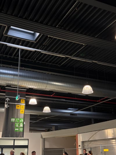 This interior shot shows the airport of Lautzenhausen, Germany. The walls are black with thin parallel lines. Large, cylindrical metal pipes run vertically, with illuminated white lights attached to them. Signage indicating directions and services, such as "WC," "rent a car," and symbols for public transportation, is visible. The top right corner shows the upper bodies of individuals.