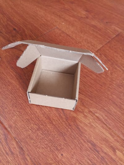 An open, empty, light brown cardboard box rests on a wooden floor. The box is box-shaped with four sides and a bottom, and its lid is hinged and folded back. The wooden floor has a reddish-brown color and a wood grain pattern with visible seams between planks. The scene appears to be indoors, and there are no people or other objects present in the image. The photograph is taken from a slightly elevated angle, looking down at the box. The lighting suggests it is daytime. The location in Vilyuysk, Russia, is not ascertainable from the image.