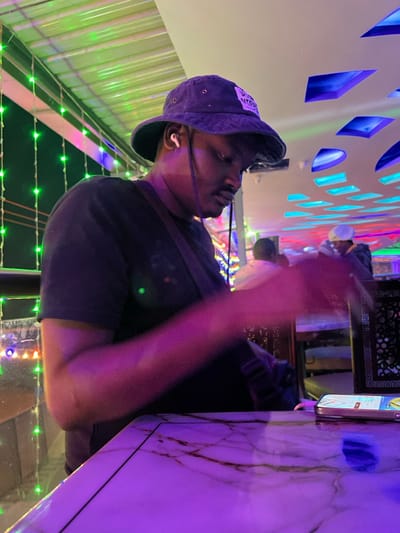 The image, captured in Kimorori, Kenya, features a man in an indoor or semi-outdoor setting, likely a lounge or restaurant, illuminated by vibrant artificial lights at night. The man is viewed from a low angle, looking upwards and to his left. He wears a dark bucket hat, which has a white patch on the front with partially visible dark text including "YOU" (possibly part of "MY YOU" or "DO YOU"). White wireless earbuds are in his ears. He is dressed in a dark t-shirt, and a dark strap, possibly from a bag, crosses his shoulder. His right arm is blurred, indicating motion, perhaps reaching or gesturing.
To the right of the man, a white table with dark, marble-like veins is visible, upon which a smartphone rests. The background showcases elaborate lighting. Above the man, a wall or ceiling features geometric cut-out shapes illuminated in blue and purple. Below him, a structure or ground area is decorated with green string lights. The overall ambiance is dynamic and modern, dominated by the glow of purple, blue, and green lights. The man's expression is difficult to discern due to the angle and dramatic lighting, but he appears focused.