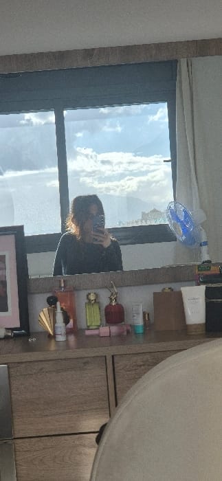 The image displays an indoor scene in Tixeraïne, Algeria, captured as a selfie reflected in a mirror. A person, identified as female, with light brown hair pulled back, is visible from the chest up, wearing a dark long-sleeved garment. She holds a smartphone to her face, obscuring it, as she takes the photograph in the mirror.

The mirror is mounted above a light-colored wooden dresser featuring two visible drawers. On the dresser surface, various objects are arranged: multiple perfume bottles of different colors (gold, green, red) and designs, a white pump bottle, a golden fan-shaped decorative item, small pink cylindrical containers, a white tube of "CLINIQUE" branded lotion, and a dark rectangular object. To the far left, a black-framed picture or mirror with a pink mat is present. To the right, a small blue and white electric fan with visibly rotating blades is positioned on the dresser.

Behind the person, through a multi-paned window with dark frames, an outdoor view is discernible. The sky is partly cloudy with blue patches. Distant buildings and subtle mountainous terrain are visible on the horizon. A light-colored curtain partially covers the window on the right side.