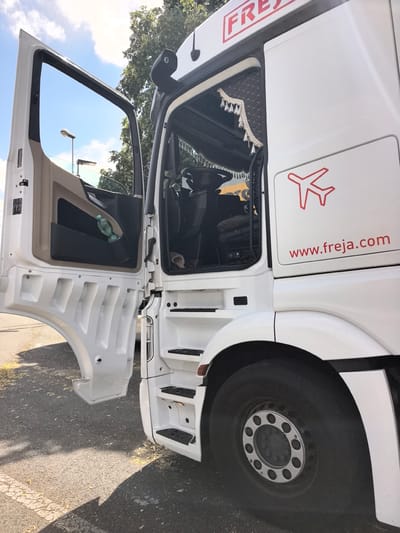 The image shows the side view of a white semi-truck with its driver's side door open. The truck is parked on an asphalt surface, possibly a road or parking lot, with some fallen leaves scattered around. In the background, there are trees and a blue sky with some clouds, suggesting it is daytime and the weather is fair.
Visible on the truck are several details: a red logo resembling an airplane and the text "FREJA" in a red box on the upper part of the cab. On the side of the truck, there's another red airplane logo and the website address "www.freja.com". Inside the open door, a plastic bottle is visible in a door pocket. The steps leading into the truck are also visible, with anti-slip treads. The truck appears to be a commercial vehicle, likely used for transportation. There are no people visible in the image, and the scene is static, suggesting the truck is parked. The overall impression is one of a professional vehicle on a bright, sunny day.