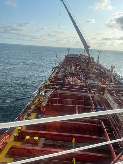 An elevated view captures the port side of a large, red-hulled tanker engaged in an offshore operation near Orimedu, Nigeria. The vessel's deck is densely populated with a network of red and yellow-piped cargo lines, valves, and various deck equipment. A prominent crane with its boom extended is positioned mid-ship. A large black hose extends from the ship's port side, connecting to a long, floating boom system in the blue-green ocean water. One individual, wearing blue-green workwear, is visible on the port side deck, near a yellow-railed platform, observing or attending to the hose connection point. Yellow bollards and safety markings, including a "SAFETY FIRST" sign, are visible on deck. The sky is partly cloudy with blue patches, and the sea exhibits moderate wave activity.