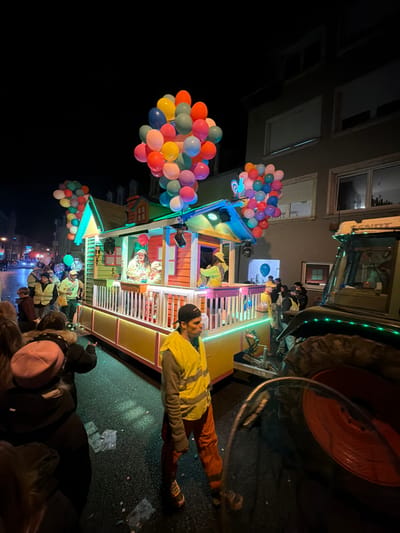A nighttime scene depicts a parade float on a street in Wasserbillig, Luxembourg. The float, designed as a small, brightly lit house with a green-trimmed roof and yellow base, is adorned with large clusters of multi-colored balloons in red, orange, yellow, green, blue, pink, and purple. Green LED strips illuminate the float's exterior. Several individuals are on the float, including one in a white top and red hat in the center and another in yellow on the right, interacting. The float is connected to a green Fendt tractor, partially visible on the far right. On the street, a male individual in a grey shirt, orange pants, yellow reflective vest, and dark baseball cap walks in the foreground. Spectators and other participants, some wearing yellow reflective vests, line the left side of the street. The asphalt road shows scattered litter. Multi-story buildings with illuminated windows are visible in the dark background.