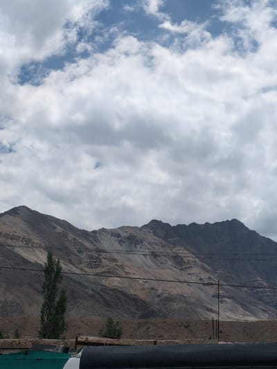 The image shows a mountainous landscape under a cloudy sky. The mountains are barren and appear to be made of rock and soil with various shades of brown and grey. There are some sparse trees and vegetation at the base of the mountains. In the foreground, there is a low wall or structure with some dark cylindrical objects on top, and a green surface to the left. Several power lines are visible across the scene, stretching from left to right. The sky is filled with white and grey clouds, with patches of blue sky visible. The overall impression is one of a rugged, natural environment, likely in a high-altitude region. The location is identified as Nimo, India.