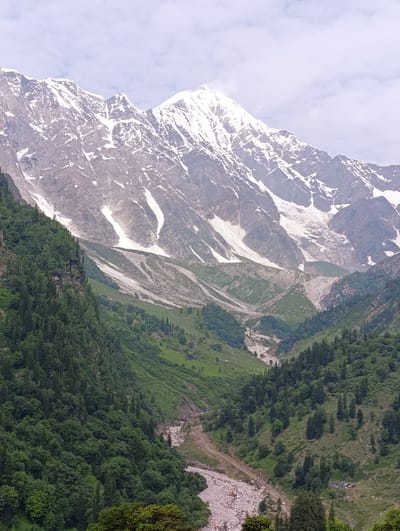 This is a vertical, eye-level, full shot photograph of a mountain valley in Dhundi, India. The upper portion of the image is dominated by a snow-capped mountain range. The snow appears to be melting and has formed streaks and patches on the rocky slopes, suggesting a glacial landscape. Below the snow-capped peaks, the mountainside transitions into green slopes covered in dense vegetation, primarily trees and shrubs.

A valley carved by a river runs through the center of the image. The riverbed is filled with rocks and appears to be dry or with low water flow, indicating it might be a seasonal river or a glacial meltwater stream. The slopes on either side of the valley are covered with lush green forests, with a mix of deciduous and coniferous trees. In the lower right corner of the image, there are a few small structures, possibly tents or cabins, clustered together, suggesting a campsite or a small settlement. The sky above is overcast with light clouds. The overall impression is a vast, rugged, and natural landscape with evidence of human presence in a small encampment within the valley.