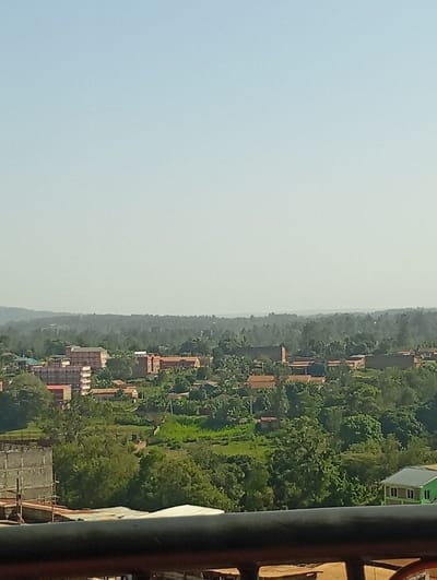 This image captures an elevated, panoramic view of a section of Murang'a town, Kenya, under a clear, hazy sky. The foreground is partially obscured by a dark, horizontal railing or ledge, indicating the perspective is from a high vantage point such as a balcony or rooftop.

The midground displays a mixed urban and natural landscape. Numerous buildings of varying sizes and styles are interspersed with dense green trees and patches of lighter green vegetation. On the left side, a multi-story building is under construction, identifiable by its exposed concrete framework. Adjacent and further into the midground, several established multi-story buildings with light-colored facades and reddish-brown roofs are visible. Throughout the scene, other structures with reddish-brown tiled or corrugated iron roofs are dispersed among the foliage. Towards the right, a distinctive building with a light green roof and light-colored walls is present. No individuals, explicit actions, or interactions are discernible within the frame.

The background consists of a distant, hazy mountain range or hills stretching across the horizon, beneath a uniformly light blue-grey sky. The overall environment suggests a developing urban area integrated into a lush, hilly terrain.