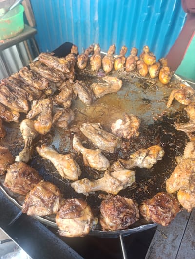 The image displays an outdoor food preparation or vending scene in Kahawa Wendani ward, Kenya. A large, circular, heavily used metal griddle, approximately 30-36 inches in diameter, is filled with numerous pieces of cooked chicken. The griddle's surface is dark, marked by congealed grease, oil, and charred residue. Approximately 50-70 chicken pieces, including drumsticks, thighs, and wings, are visible, exhibiting varied levels of browning from golden-brown to dark, almost charred. A distinct horizontal row of nine golden-brown drumsticks is positioned at the upper right section of the griddle. A partial view of a person's arm, with dark skin and a red-striped sleeve, enters the frame from the right, positioned near the chicken, indicating interaction with the food. The background consists of a corrugated metal sheet wall, painted a faded light blue. To the far left, a bright green plastic basin containing reddish material, possibly chopped tomatoes or a condiment, is visible on a simple stand. The scene is brightly lit, suggesting daylight.