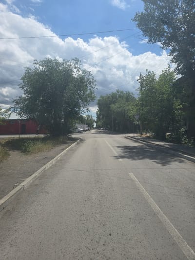The image depicts a street scene in Saran, Kazakhstan, under a partly cloudy sky. A paved road with a single white lane line runs through the center, leading towards a distant intersection. On the left side of the road, a gravel shoulder is bordered by a concrete curb and overgrown grass. Beyond the shoulder, a large, mature tree shades a section of the ground, and a red industrial-style building is visible. Further down the road on the left, a white car is parked.
The right side of the road features a sidewalk with a concrete curb, also lined with trees. A triangular yield sign is posted on the right, directing traffic. Shadows from the trees are cast across the road. The background reveals more trees and buildings, suggesting a residential or mixed-use area within the city. Utility lines are visible crisscrossing the sky. No people or vehicles are actively engaged in any actions within the frame. The overall impression is one of a quiet, everyday street in a provincial city.