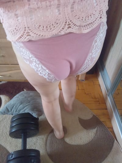 A person is standing in a room with wooden floors and a patterned carpet. The person is wearing pink lace trim underwear and a pink lace top. A dumbbell is on the carpet next to a grey pillow. A dresser is visible in the background. The image is taken from a low angle, looking up at the person. The scene is indoors and does not contain any identifiable landmarks of Terebovlia, Ukraine.