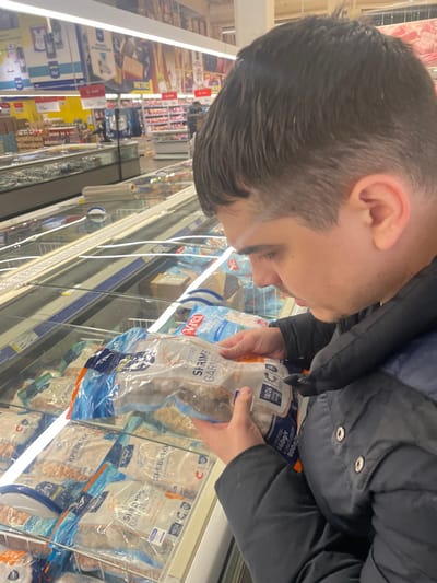 The image captures a candid moment inside a well-lit supermarket or grocery store, specifically in the frozen food aisle. The image is rotated 90 degrees counter-clockwise.
In the foreground, a person is seen from the side, facing downwards and slightly to the right (if the image were upright). They have dark hair, styled short on the sides with a fade or taper, and longer on top. They are wearing a dark, possibly black or navy blue, puffy winter jacket. Both of their hands are holding a clear plastic package of frozen shrimp, which they are intently examining.
The package of frozen shrimp has blue and white branding. Visible text on the package includes "TIGER SHRIMPS GARNELEN" and "Chef", indicating the type of product. Other details on the package show numbers like "18/26" (likely referring to the size or count of shrimp per unit of weight) and "800g/r" (indicating a weight of 800 grams). The brand name "Vici" is also partially visible, along with the word "КРЕВЕТКИ," which is Russian for "shrimps."
Behind the person, the frozen food aisle stretches into the background. Numerous other similar packages of frozen shrimp and other frozen goods are stacked inside the open-top freezers. The freezers have glowing interior lights. In the further background, more supermarket aisles are visible, lined with shelves stocked with various products. The store is brightly illuminated by fluorescent lighting, characteristic of a retail environment. The overall impression is of a person selecting or inspecting frozen seafood during a shopping trip.