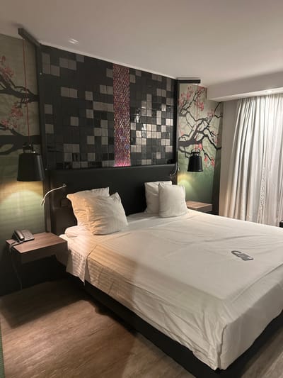 A bedroom interior is depicted, likely in a hotel in Genk, Belgium. The room features a bed with white linens and pillows, positioned against a headboard tiled with black and gray squares with a section of colorful round tiles. A wallpaper mural showing a tree branch design is visible behind the headboard, and there are black lamps mounted on either side of the bed. A bedside table is located on one side of the bed, holding a telephone. The floor appears to be wood-like. The room is illuminated, with daylight coming in through the curtains.
