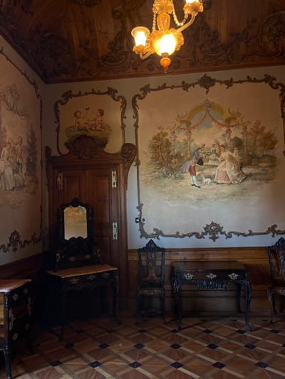 An ornate room features a high ceiling adorned with painted scenes of figures and cherubs within decorative borders. An antique chandelier hangs from the elaborately carved wooden ceiling. Dark wood furniture, including tables, chairs, and a mirror, lines the walls. The floor is patterned with geometric tiles in shades of brown.