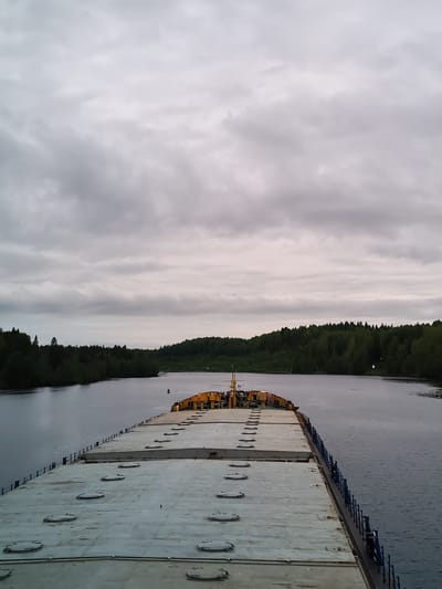 The image shows a view from the deck of a barge on a waterway near Volokov Most, Russia. The barge has a gray, weathered deck with numerous circular access covers. Blue railings line the edges of the barge. The waterway is bordered by dense green forests. The sky is overcast with heavy gray clouds. The overall scene appears calm and overcast.