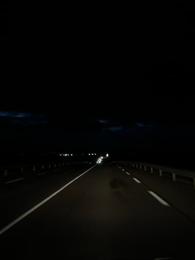 A nighttime scene from an elevated perspective, likely a bridge, dominates the image. The roadway stretches into the distance, illuminated by distant lights and lane markings. To the left, dark water reflects faint glimmers, possibly moonlight or city lights. A stark contrast divides the scene, emphasizing the dark, mysterious water and the structured, lit roadway. The composition features sharp diagonal lines and varying degrees of brightness, adding depth and visual interest to the dark, moody atmosphere.