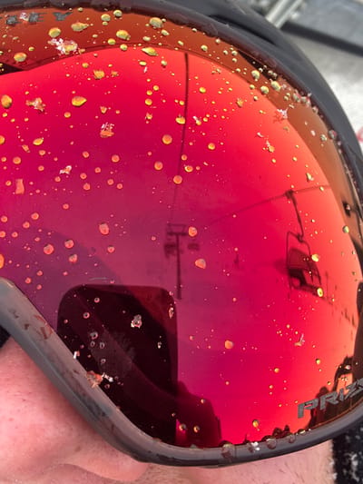 A close-up, first-person perspective photograph depicts a red mirrored ski goggle lens, covered with numerous small, irregular yellow-orange water droplets and several white ice particulates. The goggle frame, dark in color, borders the lens along the top and bottom. The brand name "PRIZM" is visible on the lower right portion of the lens. Reflected in the lens is a ski lift structure, consisting of a vertical pole and at least two unoccupied chairlift seats, against a diffuse, bright background indicative of a snowy slope or sky. A flesh-toned area, consistent with a person's chin or cheek, is reflected in the lower-left quadrant, alongside a dark, angular reflection in the lower-center. The environment is a snowy, outdoor mountain setting in St. Christoph, Austria, typical of a ski resort.