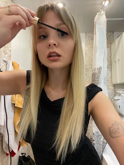 A young Caucasian female with long blonde hair and bangs is depicted in a bathroom, applying mascara to her right eye using a black-bristled wand with a gold handle held in her left hand. She wears a black textured top, a gold ring with a green stone on her left ring finger, a thin gold necklace, and a small gold earring on her right ear. An ornate, light-colored, heart-shaped tattoo is visible on her right upper arm. The background features light-toned patterned wall tiles, a yellow and white towel hanging on the left, a partially visible white shower curtain with a brown pattern on the right, and a white two-door cabinet in the upper right. A crumpled black plastic bag is present on the lower left. Two overhead light fixtures illuminate the scene. This scene occurs in a bathroom in the city of Vysokovo, Russia.