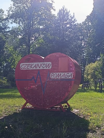 The image shows a large, red, heart-shaped sculpture in Czekanów, Poland. The sculpture is made of metal mesh and has text on it. On the left side of the heart, the word "CZEKANOW" is written in white letters with a small heart replacing the "A". Below this text, a blue electrocardiogram (ECG) line is depicted. On the right side of the heart, the word "POMAGA" is written in white letters. The sculpture is supported by a red metal stand and is situated outdoors on a grassy area with trees in the background. Sunlight is filtering through the trees, creating a bright and somewhat overexposed effect in some parts of the image. The overall scene appears to be a park or public space.