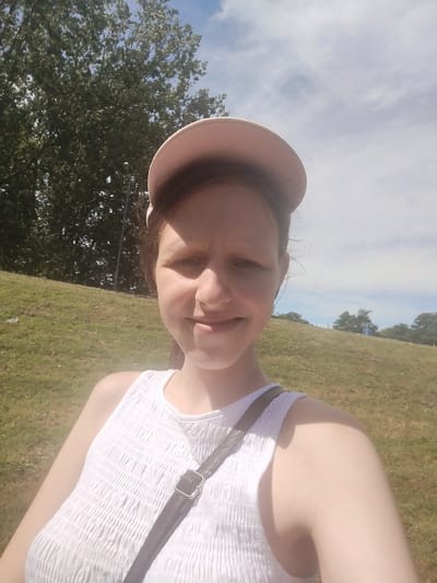 A young white female wearing a pink baseball cap and a white sleeveless top takes a selfie outdoors. She is positioned in the foreground, looking directly at the camera with a slight smile. Her face is partially obscured by the brim of her cap. Behind her, a grassy hill slopes upwards, with trees visible in the background against a cloudy blue sky. A dark strap from a bag crosses diagonally over her chest. The lighting suggests it is daytime, likely during warm weather. The overall setting appears to be a park or an outdoor recreational area, possibly in the city of Cwmbran, United Kingdom, given the presence of well-maintained greenery and open spaces.