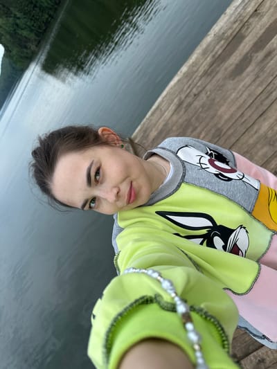 A young woman with brown hair, green eyes, and freckles is taking a selfie on a wooden pier by a body of water. She is wearing a multi-colored sweatshirt with cartoon characters on it. The water is calm and reflects the surrounding trees, which are lush and green. The pier is made of weathered wood planks. The scene appears to be in a natural, outdoor setting, possibly a lake or reservoir, and the mention of Zhebrei, Russia, provides a geographical context for the location.