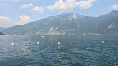 This is an eye-level, medium shot of Lake Garda, Italy, with a view of the mountains behind Limone sul Garda. In the foreground, the deep blue water is calm with ripples. Several white buoys are scattered across the lake, marking a section of the water. In the mid-ground, a small white boat with a red stripe and one person wearing a red shirt is visible on the left side of the frame. Further to the right, another white boat is moving. In the background, the imposing, green-covered mountains rise dramatically, their rocky faces interspersed with lighter patches. A small, densely populated town is nestled at the base of the mountains on the right side of the frame. The sky above is a pale blue with scattered white clouds. A faint speck, possibly a bird, is visible in the sky above the mountains.