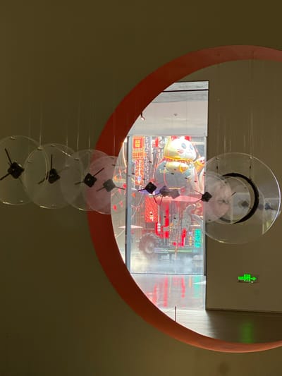 In the Jinniu District of China, an indoor art installation features a large, orange, circular frame partially obscuring a display visible through a glass window. Suspended from the ceiling are several clear, circular discs, resembling clock faces, each with black hour and minute hands. The discs are arranged in a linear fashion.
Behind the discs and within the circular frame, the window reveals a vibrant and eclectic display. Dominating the scene is a large, cartoonish character, possibly a cat or bear, adorned in a blue and gold outfit, sitting atop a complex, industrial-looking structure. This structure appears to be a vehicle, possibly a tractor, with large wheels and intricate mechanical details. The display is illuminated by bright lights, creating a colorful and somewhat chaotic aesthetic. Red and green neon signs with Chinese characters are visible within the display, adding to the visual intensity. The floor in front of the display is wet, with reflections of the lights and signage creating a shimmering effect. To the right of the display, a green exit sign with Chinese characters is mounted on a plain, light-colored wall. The overall environment suggests an exhibition space or a gallery.