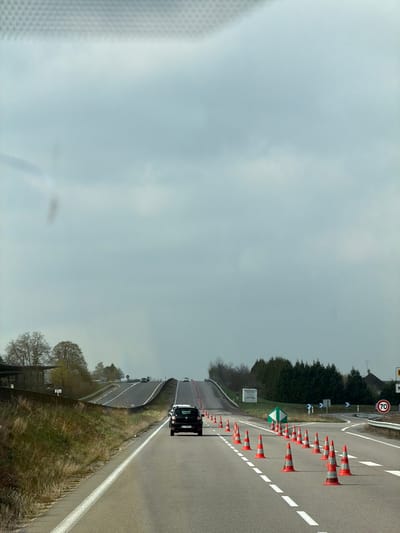 The image depicts a multi-lane road scene in Pagny-sur-Meuse, France, viewed from inside a vehicle's front passenger compartment. The sky is overcast with a light grey cloud cover. The primary subject is a paved asphalt road extending into the distance, ascending a gentle incline.
In the foreground and middle ground, the road features white dashed and solid lane markings. Directly ahead, in the right lane, a dark-colored Dacia Duster SUV is proceeding forward. To the right of the Duster, a long line of orange and white traffic cones marks a temporary lane closure or redirection, guiding traffic towards the right. This cone-delineated path leads to an intersection or diverging road branch, where a circular white speed limit sign indicating "70" is visible on the right shoulder, alongside a green diamond-shaped sign with white arrows indicating a split in directions.
On the left side of the main road, a grassy embankment borders a separate, elevated road or overpass that also ascends into the distance. Several indistinct vehicles are visible on this upper road and in the far distance on the main thoroughfare. The immediate roadside on the left features a concrete barrier and sparse, deciduous trees. On the right, beyond the coned-off section, there are clusters of evergreen trees and a partial view of a building with a dark roof. No individuals are visible in the scene. The overall environment suggests a rural or semi-rural highway context with ongoing road adjustments.