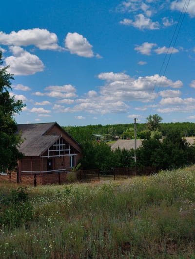 The image depicts a rural scene in Matiashivka, Ukraine, under a bright blue sky dotted with fluffy white clouds. The foreground is a field of tall, wild grasses and small white wildflowers, suggesting a warm, perhaps late spring or summer day. A weathered brown fence runs across the middle ground, separating the wildflower field from a yard where a brick building stands.

The building appears to be a house or a small community structure, with a dark grey, angled roof made of slate or corrugated metal. It has a prominent gable with a white, latticed window. A dark-colored door is visible under the porch, and a small white window is to its right. The building is surrounded by lush green trees, which also form a dense line of foliage in the background, hinting at a wooded area or a cluster of rural homes.

To the right of the building, a tall wooden pole supports electrical wires that stretch diagonally across the sky, a common sight in rural landscapes. Beyond the immediate building and trees, other rooftops and structures can be glimpsed, suggesting a small village or hamlet. The overall atmosphere is peaceful and serene, evoking a sense of rural tranquility on a sunny day. There is no visible text in the image.