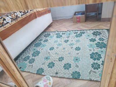A room interior is reflected in a mirror. A bed with a patterned duvet and brown trim is visible on the left. A rug with a teal and grey floral pattern covers the wooden floor. A cardboard box with items inside is on the floor in the background. A wooden chair and cabinet are also visible. A slipper with a floral pattern is on the floor in the foreground, and is also reflected in the mirror.