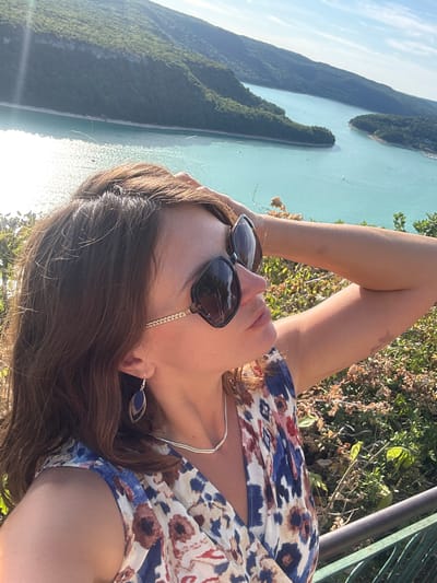 A woman is taking a selfie outdoors with a scenic body of water and forested hills in the background. She is wearing sunglasses, a silver necklace, and a floral print dress with blue, white, red, and brown patterns. Her hair is brown and she has a blue earring in her visible ear. She has her right arm behind her head, and her left arm is resting on her body. The background shows a large, turquoise lake with several boats, surrounded by lush green hills. The sun is shining brightly, creating a glare on the water and casting shadows. The image appears to be taken from a vantage point overlooking the lake. The location is identified as Moirans-en-Montagne, France.