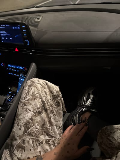 The image captures a close-up view inside a car, likely at night or in a dimly lit environment, from an elevated perspective looking down towards the passenger seat area.
In the center and right side of the frame, the right leg, foot, and hand of a person are visible. The person is wearing a black athletic shoe with distinct silver or metallic grey accents and white laces. Dark-colored leggings or pants are worn on the leg. Resting on the person's lap or thigh is a piece of fabric, possibly a blanket or part of an outfit, featuring a camouflage or abstract mottled pattern in shades of light grey, white, tan, and dark brown/green.
The person's right hand is positioned on or near their leg. The fingernails are long, pointed, and appear to be painted a dark color, possibly black. The back of the hand and wrist are extensively tattooed with numerous small, scattered star and geometric shapes, along with a larger, darker tattoo partially visible further up the arm.
The left and lower parts of the image reveal sections of the car's interior. A digital infotainment screen is visible on the dashboard, displaying information with a blue backlight. Text on the screen includes "10:36," which could indicate time or a music track duration. Faintly visible song or artist names might include "SHAD" and "MARO". Below and to the right of the screen, various car controls and buttons are present, some illuminated with a blue glow. A prominent red triangular button, likely for hazard lights, is also visible. The dashboard and door panel surfaces feature a dark, textured material, possibly a carbon fiber-like pattern. The overall lighting is dim, indicating it is dark outside, with the car's internal lights providing the main illumination.