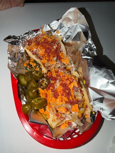 The image shows a red basket lined with aluminum foil containing a Tampico-style sandwich. The sandwich is topped with shredded cheese, crumbled red meat, and pickled jalapenos. The basket is on a white surface. A portion of a hand is visible on the left side. The location is Tampico, Mexico, implied by the sandwich style.