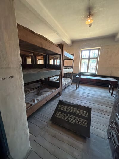 The image depicts the interior of a former barracks at the Auschwitz-Birkenau Memorial and Museum in Oświęcim, Poland. The room contains several tiered wooden bunks, each with a thin mattress covered in a light-colored fabric. The walls are bare and appear to be made of plaster. A barred window is visible on the right side of the room, letting in natural light. A simple wooden table and bench are positioned near the window. A plaque with text, written in both Polish and English, is placed on the floor in the foreground, providing historical information about the location. The overall impression is one of austerity and historical significance.
