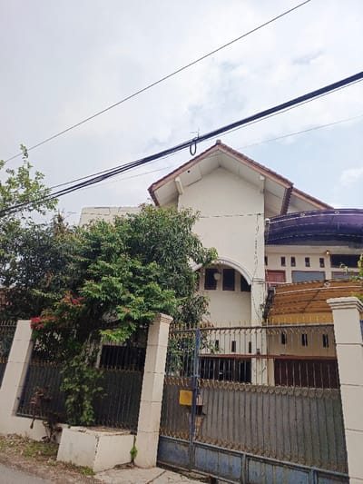A two-story house is partially obscured by a green tree and a gray metal fence with concrete posts. The house is white with a red-tiled roof. An arched feature with windows is visible. Another building with a purple awning is connected to the main house. Overhead are black electrical wires against a cloudy sky.