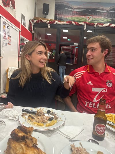 The image depicts two individuals seated at a table inside a Portuguese restaurant, likely Casa Portuguesa, located in Décines-Charpieu, France.
On the left, a young adult female with long, light brown hair, wearing a dark blue knitted sweater, is smiling broadly and looking towards the male. Her right hand is slightly raised, near the male's arm. On the right, a young adult male with short, curly brown hair wears a red Adidas polo shirt with white stripes, an SL Benfica crest, and a "TELCEL" sponsor logo. He is also smiling and looking at the female. In the background, a third male figure in a dark jacket and blue jeans is visible through a glass door.
The white-clothed table features several dishes. In front of the female is a white plate containing remnants of a meal, including shredded chicken, rice, black olives, and fried plantains/potatoes, with a knife and fork resting on it. Another plate in the foreground holds several pieces of roasted chicken. A brown glass bottle of Super Bock beer stands upright on the table to the right, next to a plate with french fries. White paper napkins and a drinking glass are also visible.
The restaurant's interior walls are white with red trim. A large banner depicting a football stadium (likely Estádio da Luz) with the SL Benfica crest is displayed on the wall behind the individuals. A red and white tinsel garland is strung across a doorway, above which a black speaker is mounted. A white sign with "SORRIA" (Smile) and a camera icon is visible on a white cabinet/door to the far left.